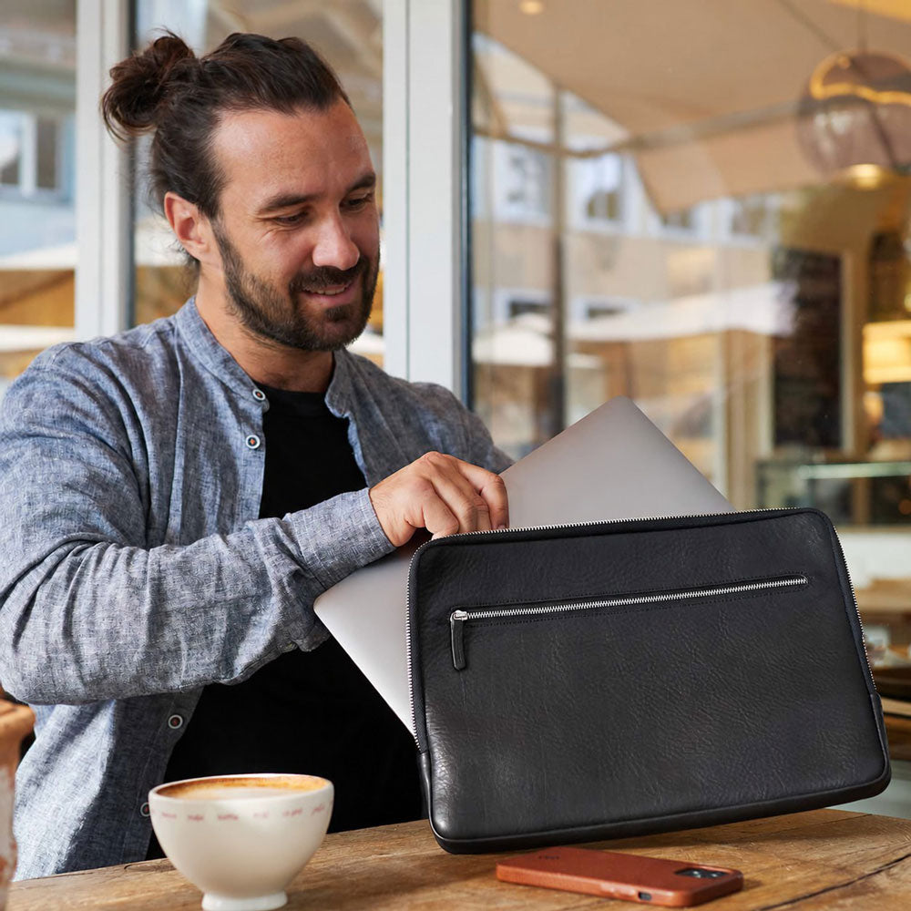 Ein Mann sitzt mit einer Macbook Pro Hülle aus Leder in Schwarz an einem Tisch und verstaut sein Macbook