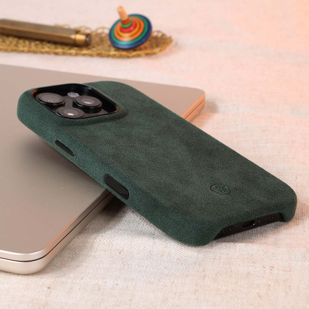 Grüne Alcantara-Hülle für iPhone 16 Pro von Good Wilhelm, handgefertigt mit integriertem Kamera-Kontrollknopf, elegant arrangiert auf einem Tisch mit Accessoires.