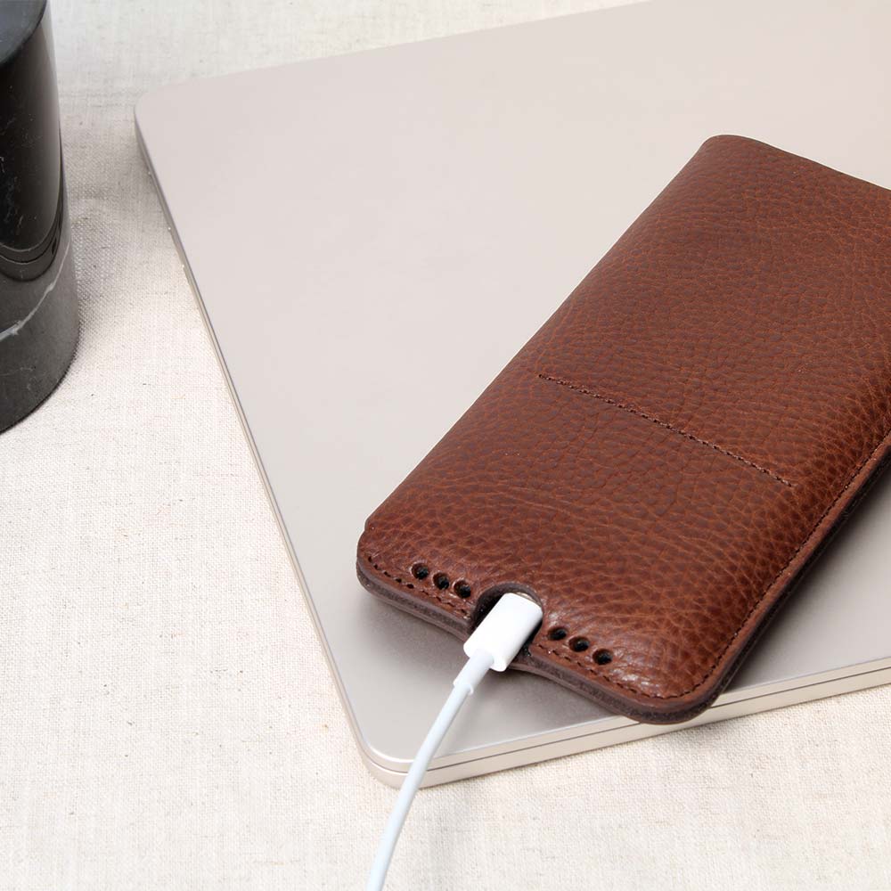 Braunes iPhone Sleeve von Good Wilhelm, aus hochwertigem Leder mit Ladeaussparung und Soundholes, angelehnt an ein MacBook auf einem Tisch – elegante Schutzhülle aus edlem Leder für optimalen Schutz und Funktionalität