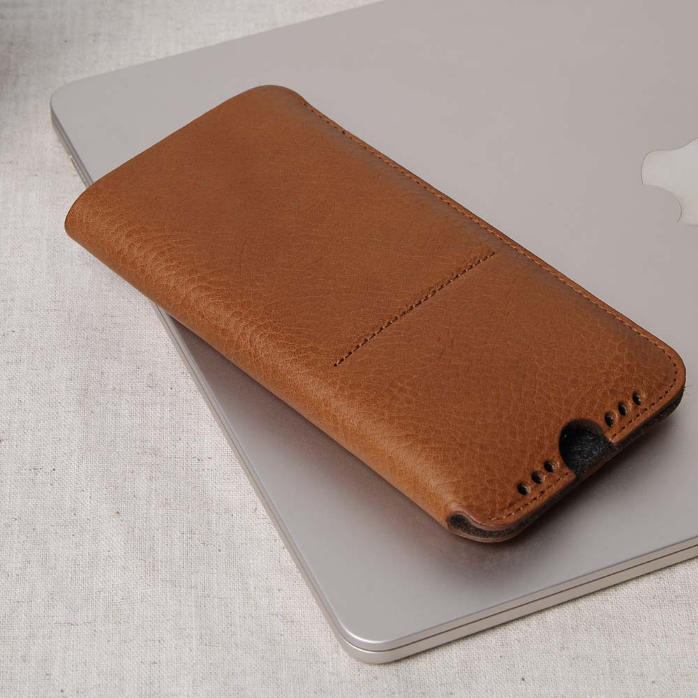 Cognac-braunes iPhone Sleeve aus hochwertigem Leder mit Ladeaussparung und Soundholes, angelehnt an ein MacBook auf einem Tisch – stilvolle Schutzhülle aus edlem Leder für optimalen Schutz und Funktionalität.